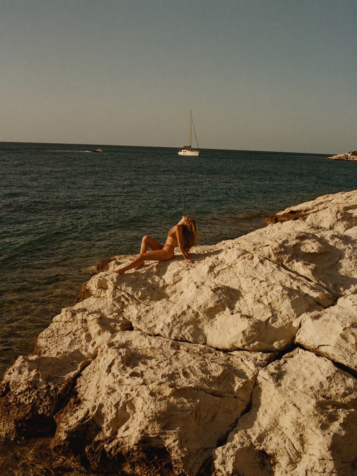 A model resting on the limestone rocks of a wide Mediterranean cove at golden hour